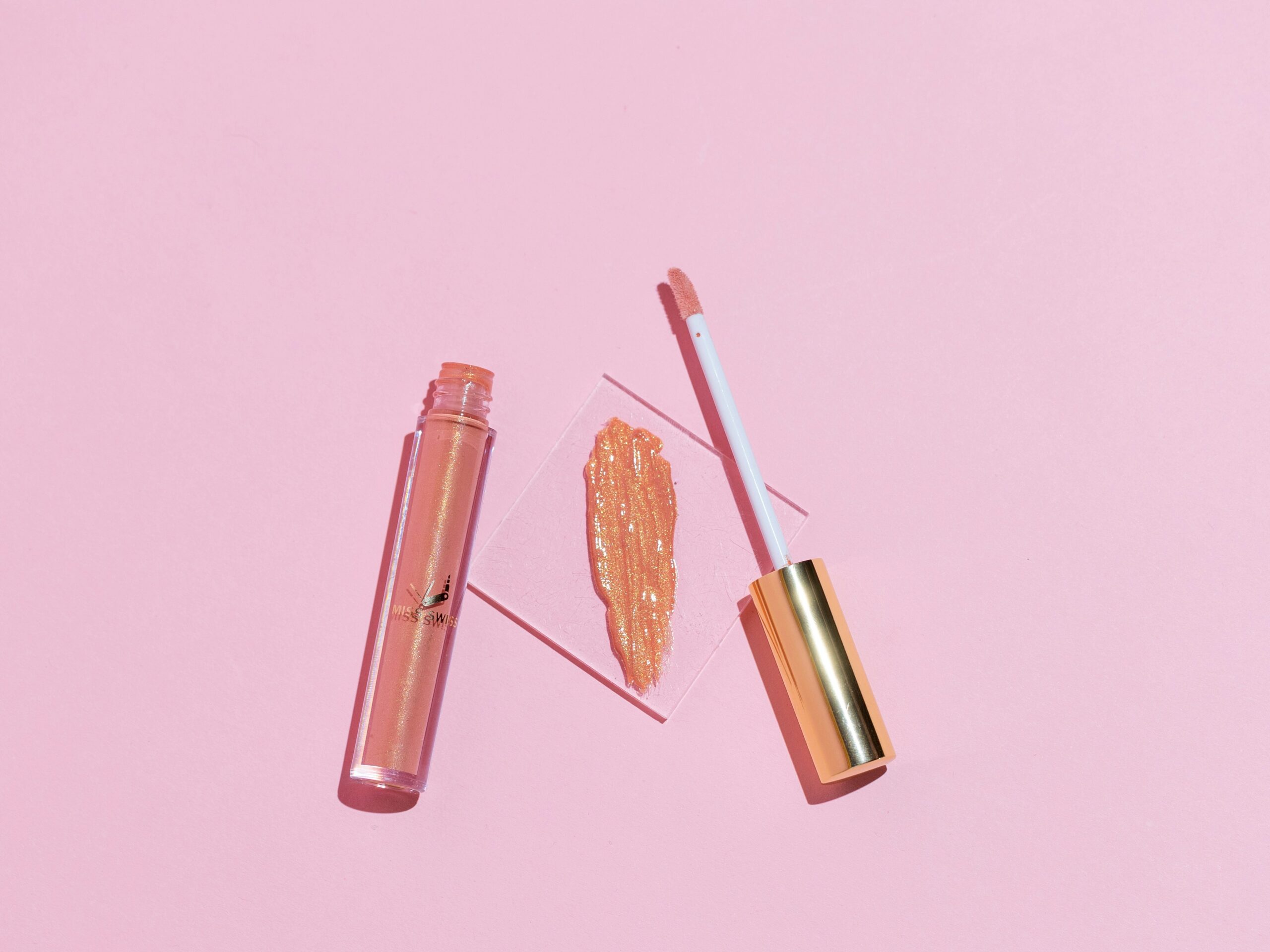Shimmering lip gloss with applicator displayed on a pink background, highlighting its glossy finish and elegant packaging.