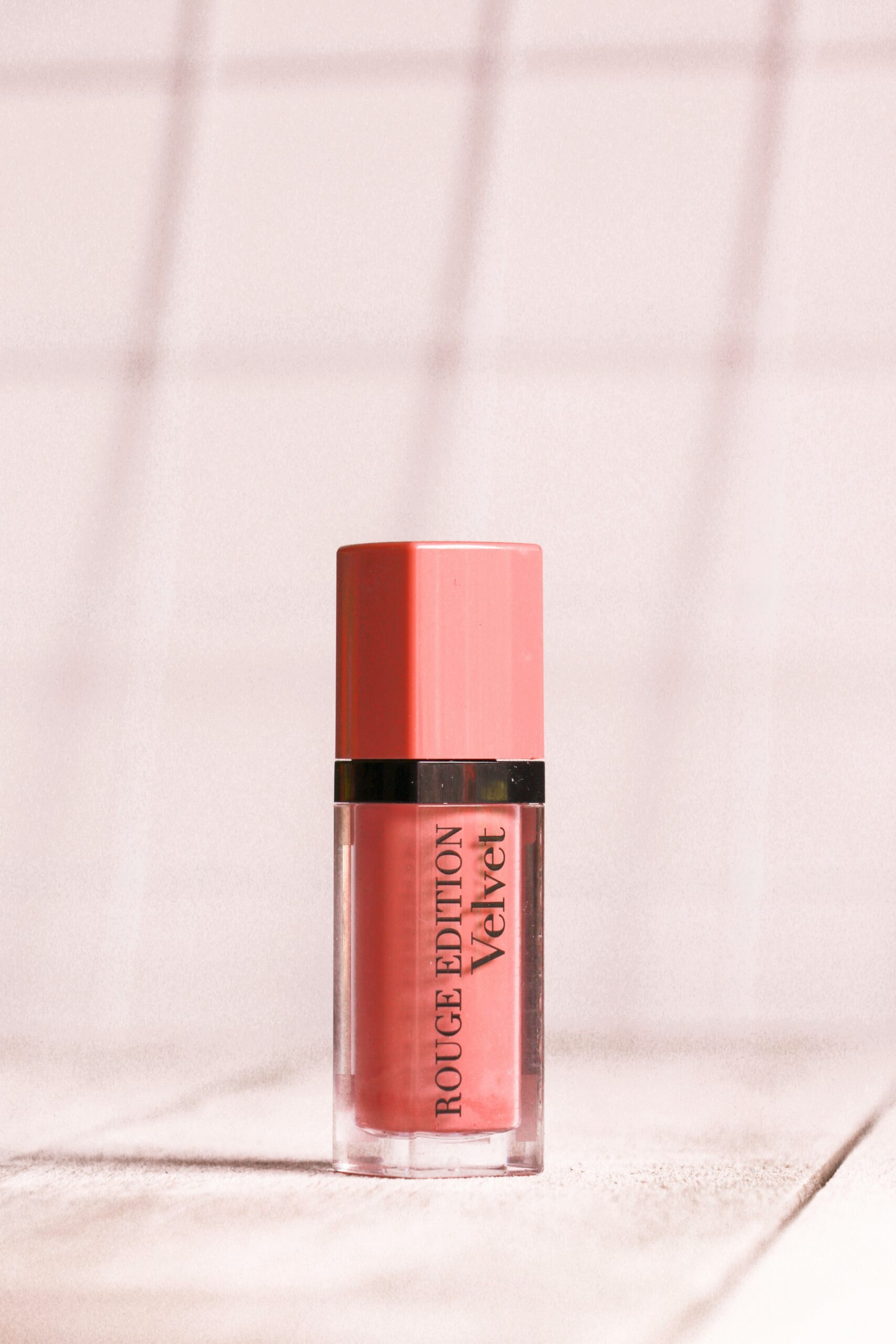 Coral pink liquid lip color in a sleek transparent tube with a matching pink cap, displayed on a soft pastel background with natural light and subtle shadows.